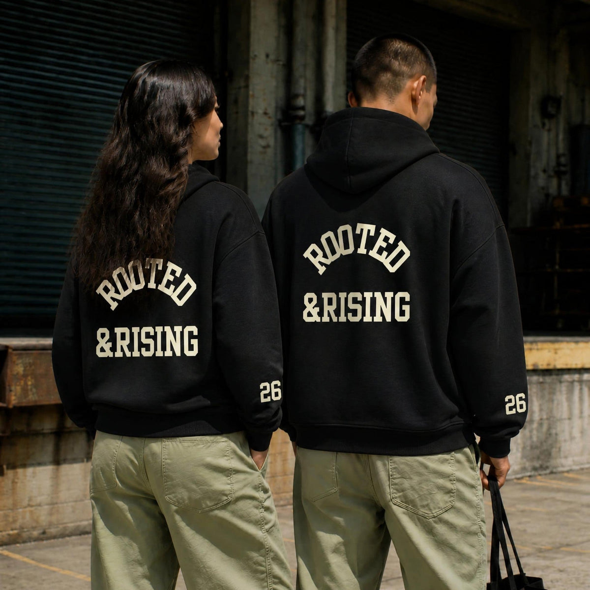 Two people wearing the Rooted & Rising Hoodie, with ROOTED &RISING varsity lettering across the back and SA 26 at the sleeves