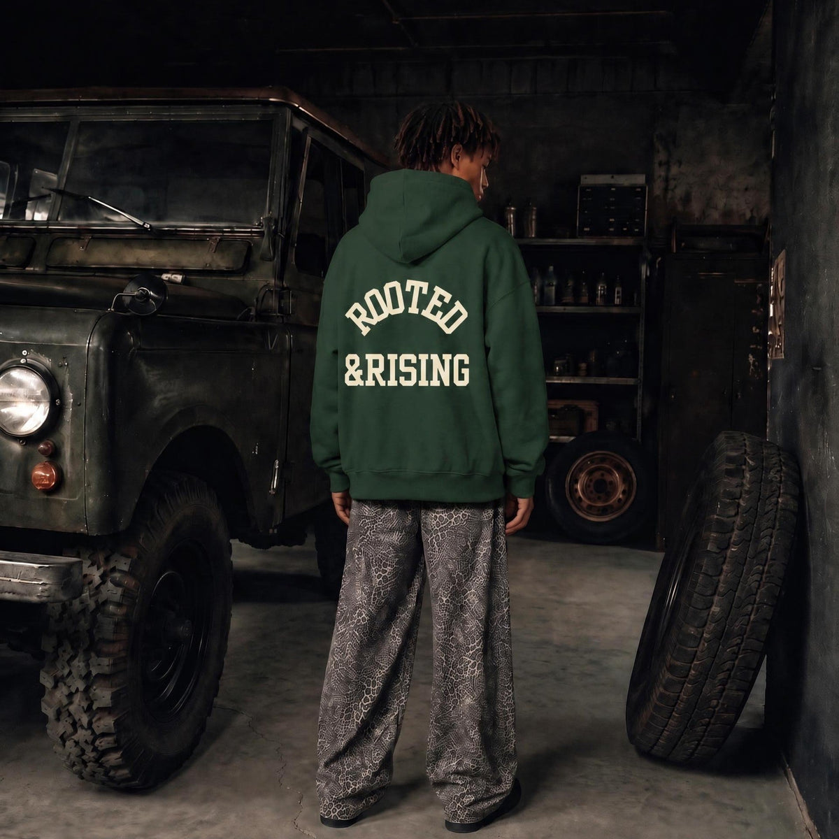 Back view of a person wearing the Rooted & Rising Zip-Through Hoodie, with ROOTED &RISING varsity lettering across the back, standing in a garage beside a vintage truck